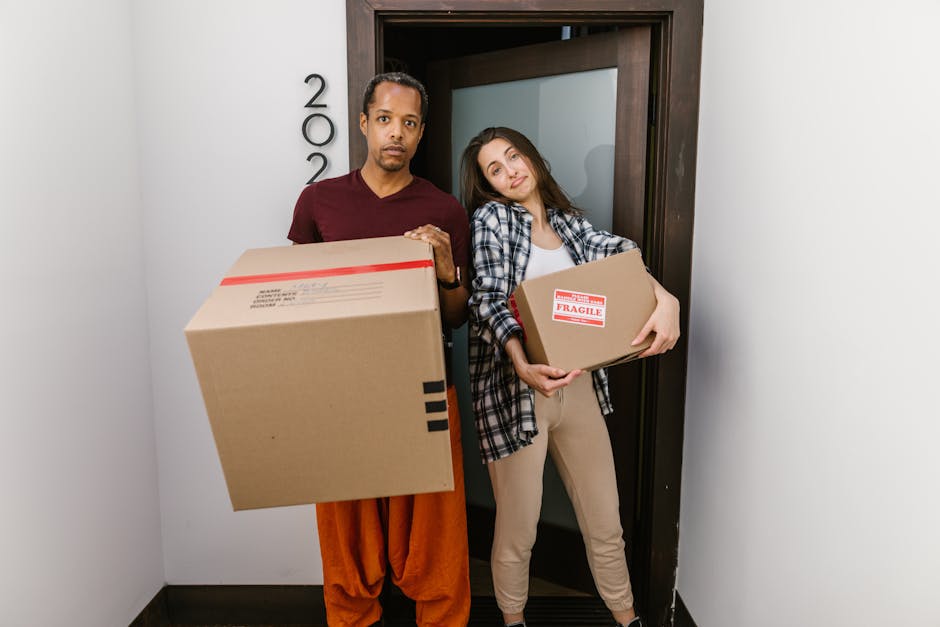 A man and a woman stand inside a doorway, each holding a cardboard box during a home relocation process. The man, on the left, has dark hair and is dressed in a maroon t-shirt and orange pants, suppor