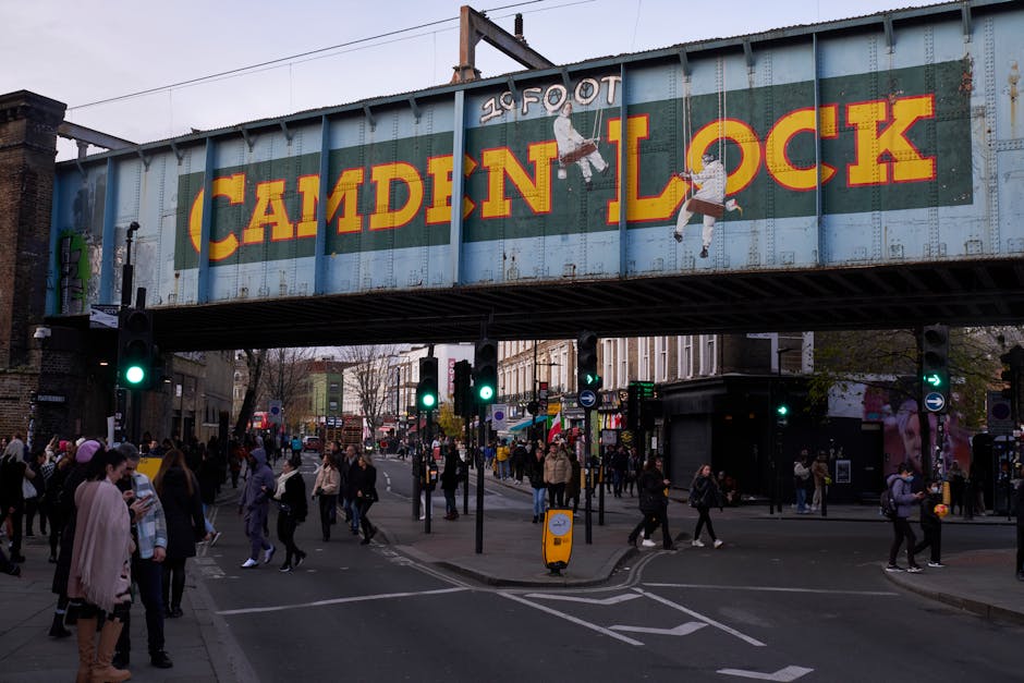 The image depicts a busy street scene under a large railway bridge with a blue and yellow 'Camden Lock' sign attached to it. Below the bridge, numerous pedestrians are crossing the road at a busy inte