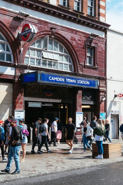 The image depicts a busy street scene under a large railway bridge with a blue and yellow 'Camden Lock' sign attached to it. Below the bridge, numerous pedestrians are crossing the road at a busy inte